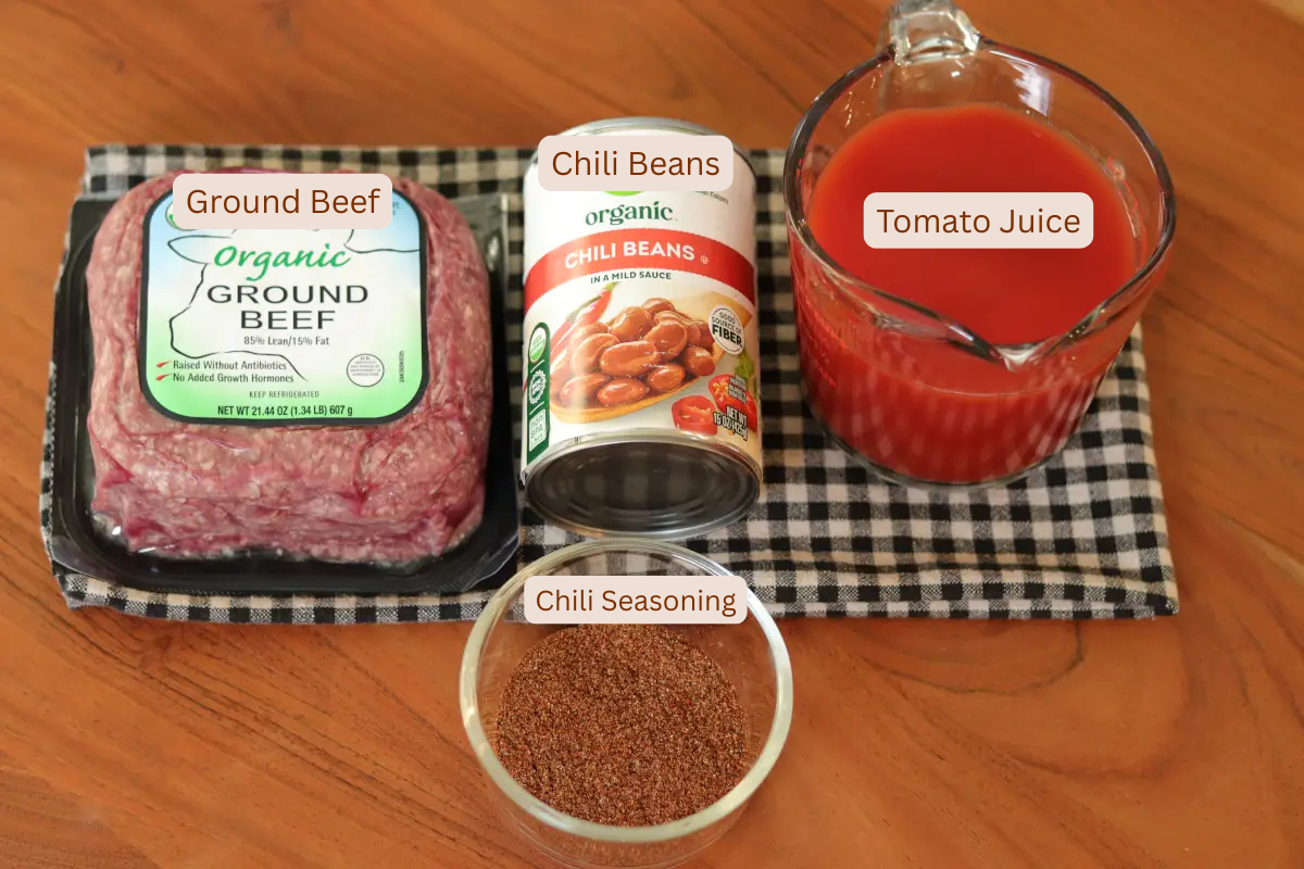 A package of ground beef, a can of chili beans, a pitcher of tomato juice, and chili seasoning on a plaid towel.
