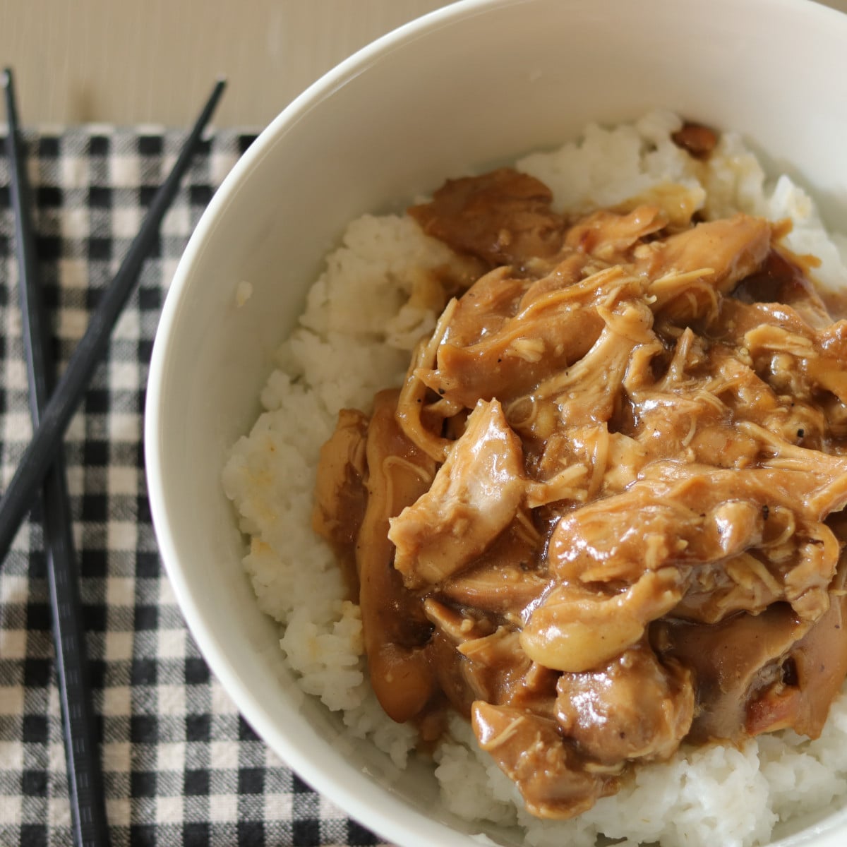 Slow cooker chicken teriyaki over a bed of rice on a plaid towel next to chopsticks.